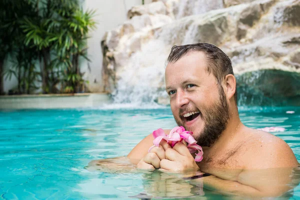 Usměvavý vousatý muž drží v bazénu květiny. Tropické květiny Frangipani Plumeria, Leelawadee plující ve vodě. Lázeňský bazén. Mír a klid. Lázeňský koncept. — Stock fotografie