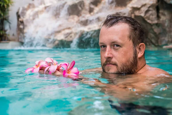 Bebaarde man houdt bloemen in het zwembad. Tropische bloemen Frangipani Plumeria, Leelawadee drijvend in het water. Spabad. Vrede en rust. Spa concept. — Stockfoto