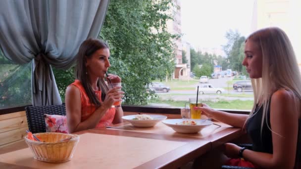 Két hosszú hajú lány pihen egy kávézóban egy modern belső és nevetve. Beltéri portréja vicces mosolygó hölgyek divatos ruhákat iszik gyümölcs smoothies. — Stock videók