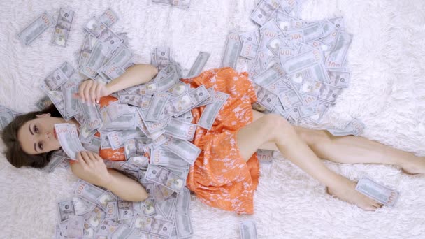 Uma menina bonita goza de uma enorme riqueza. a fêmea jaz em uma cama branca com muito dinheiro, em uma pilha de notas de dólar . — Vídeo de Stock