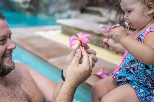 Family Spa. male and baby hands holding pink flower in turquoise pool water.