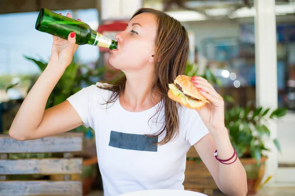 Brunette girl, with an independent look, drinks beer and eats a hamburger. The concept of feminism and female independence. Oktoberfest. — Stock Photo, Image