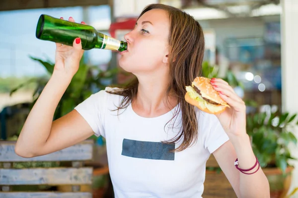 Chica morena, con un aspecto independiente, bebe cerveza y se come una hamburguesa. El concepto de feminismo e independencia femenina. Oktoberfest . — Foto de Stock