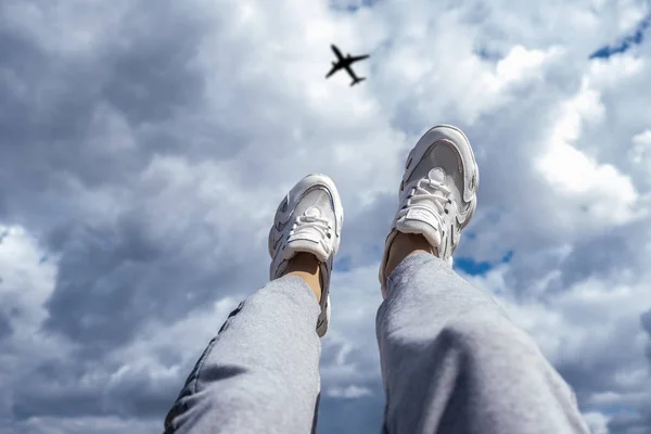 Beine in weißen Turnschuhen vor dem Hintergrund eines wolkenverhangenen Himmels und eines fliegenden Flugzeugs. Freiheitskonzept. — Stockfoto