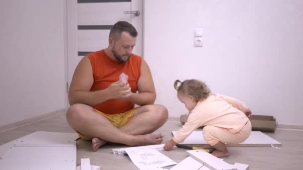 Una niña ayudando a papá a recoger muebles en una casa nueva. Un hombre recoge un gabinete de madera blanca — Vídeos de Stock