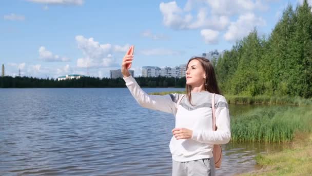 Menina bonita com uma mochila leva uma selfie perto do lago e fala em um telefone celular no Skype . — Vídeo de Stock