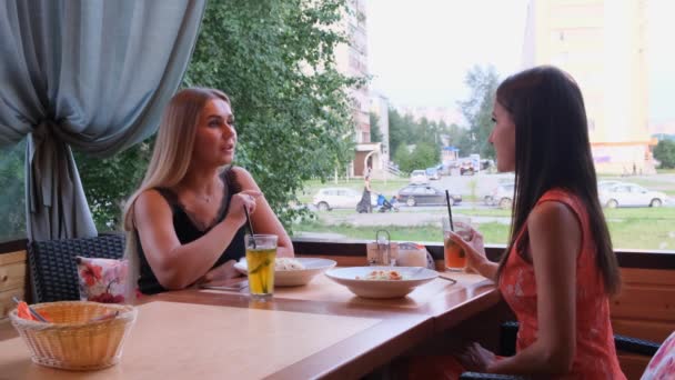 Due ragazze dai capelli lunghi stanno riposando in un caffè con un interno moderno e ridendo. Ritratto al coperto di divertenti signore sorridenti in vestiti alla moda bere frullati di frutta . — Video Stock