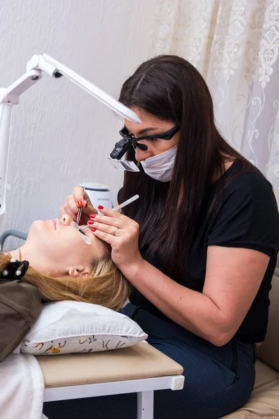 Woman Eye with Long Eyelashes. Eyelash Extension. Lashes, close up, selected focus. Gluing artificial eyelashes with tweezers. Beautician in magnifier glasses performs laborious small work.