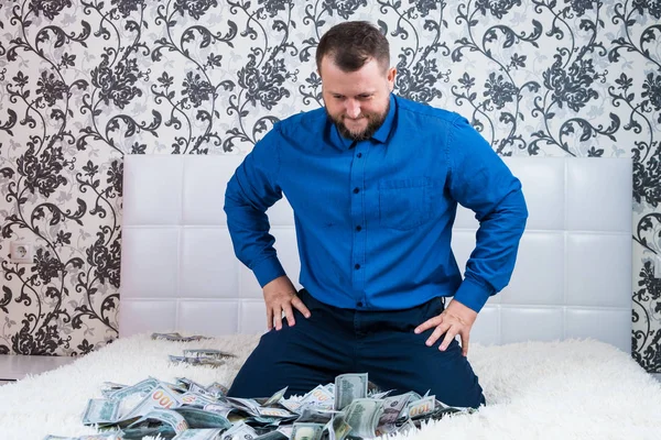 Uomo barbuto in camicia blu gode di un sacco di soldi su un letto bianco, un sacco di soldi vola in aria, un sacco di banconote di dollari . — Foto Stock