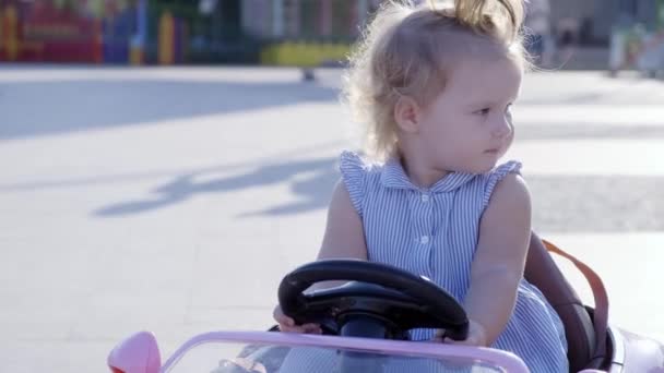 Nizhnevartovsk, Russia - August 12, 2019: little girl in a blue dress rides a pink baby car in a park. Happy childhood concept. Caricature of an adult woman driving a car. — Stock Video