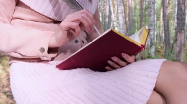 Sonbahar ormanında bir kızın yakın çekim, bir kadın el bir kitapta bir kalem ile yazıyor. Bir kadın sonbahar ormanında bir ağacın yanında oturur ve elinde bir kitap tutar.