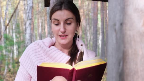 Brunette meisje in het herfst bos, leest een boek, een vrouw zit in de buurt van een boom in het herfst bos en houdt een boek in haar handen — Stockvideo