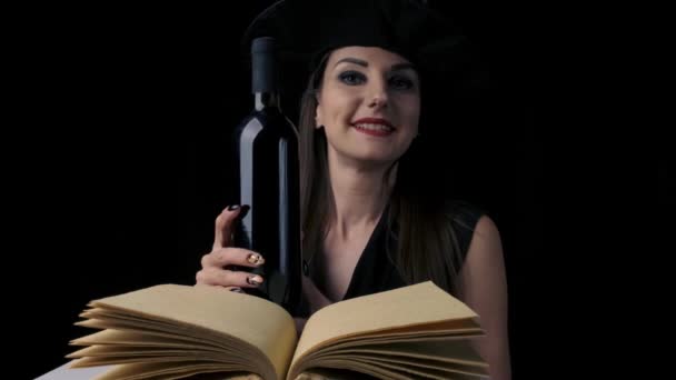 Seksowna kobieta w czarnym kostiumie czarownicy i kapeluszu, trzyma butelkę czerwonego wina w rękach, patrzy w kamerę i uśmiecha się. zbliżenie. Halloween przyjęcie świąteczne. — Wideo stockowe
