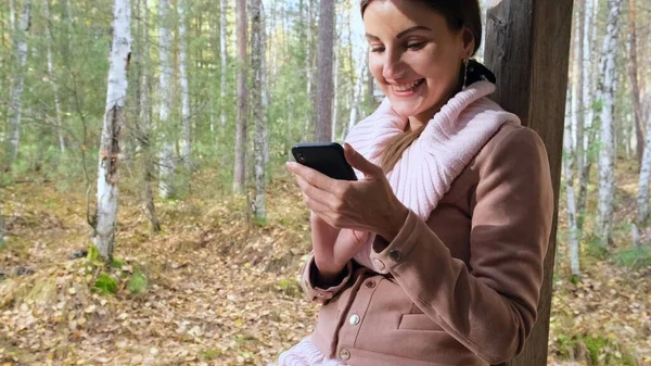 Zamyślony Kobieta w lesie jesienią rozmawiać przez telefon, wysyła wiadomości na smartfonie, czyta coś na telefon komórkowy — Zdjęcie stockowe