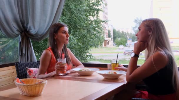 Две длинноволосые девушки отдыхают в кафе с современным интерьером и смеются. Крытый портрет смешных улыбающихся дам в модной одежде, пьющих фруктовые коктейли . — стоковое видео