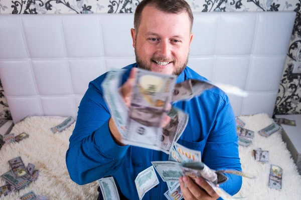 A man in a blue shirt rejoices at the tremendous wealth on the white bed, a lot of money flies in the air. — Stock Photo, Image