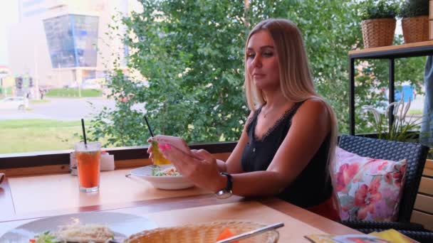 Vakker, blond kvinne som prater i telefonen, snakker i skype og drikker cocktail, sitter på en kafe på gata . – stockvideo