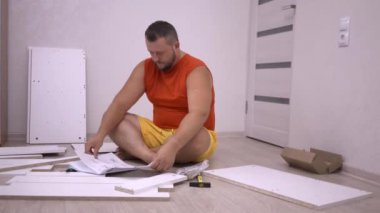 Erkek, talimatları kullanarak yeni bir evde beyaz ahşap mobilya topluyor. Bir adam kendi evinde bir dolap toplar..