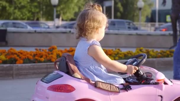 Nizhnevartovsk, Ryssland-12 augusti 2019: liten flicka i en blå klänning rider en rosa babybil i en Park. Happy Childhood koncept. Karikatyr av en vuxen kvinna som kör bil. — Stockvideo