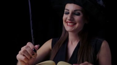 Siyah cadı kostümlü ve şapkalı güzel seksi bir kadın kalemle bir kitabı büyülüyor ve bir kitaba kalem yazıyor. Cadılar Bayramı partisi.