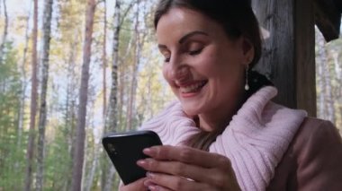 Sonbahar ormanında neşeli bir kadın telefonla konuşuyor, akıllı telefondan mesajlar gönderiyor, cep telefonundan bir şeyler okuyor.
