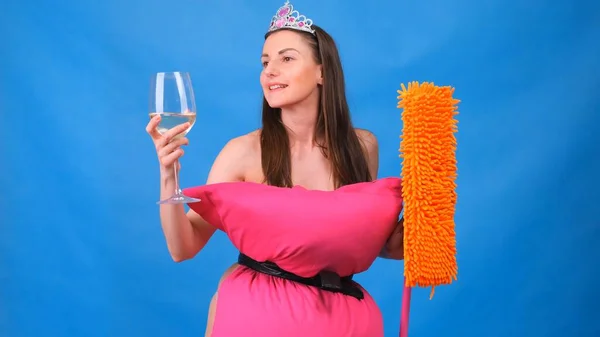 Όμορφο κορίτσι με ροζ φόρεμα από μαξιλάρια με σφουγγαρίστρα και ένα ποτήρι κρασί σε μπλε φόντο. Τρελή καραντίνα. Αστείο καθάρισμα του διαμερίσματος. Μόδα 2020. Βάλε ένα μαξιλάρι. Πρόκληση 2020 λόγω — Φωτογραφία Αρχείου
