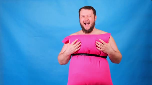 Morsom skjeggete fyr i rosa kjole laget av puter berører brystet hans på blå bakgrunn. Sprø karantene. Rart husrengjøring. Mote 2020. Legg på deg en pute. Utfordring 2020 på grunn av boligisolering – stockvideo