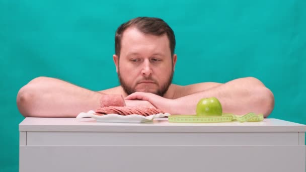 Jeune homme gras attrayant en t-shirt décontracté fait un choix facile entre la pomme verte ou la saucisse, choisissant des fruits biologiques sains au lieu de la restauration rapide malsaine, fermant les yeux et souriant joyeusement — Video