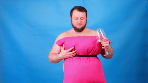 Rolig skäggig kille i rosa klänning gjord av kuddar dricker vin från ett glas på en blå bakgrund. Galen karantän. Mode 2020. Sätt på dig en kudde. Utmaning 2020 på grund av isolering — Stockvideo