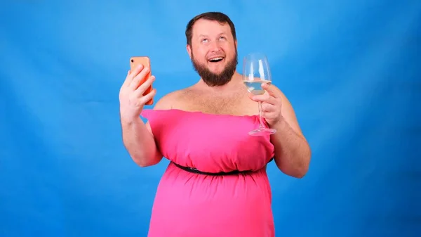 Lustiger bärtiger Typ in einem rosafarbenen Kleid aus Kissen mit einem Glas Wein macht Selfie auf blauem Hintergrund. Verrückte Quarantäne. Lustige Hausreinigung. Mode 2020. Auf ein Kissen legen. Herausforderung 2020 — Stockfoto