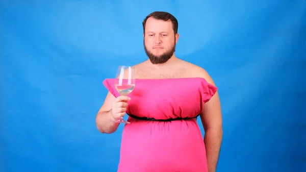 Забавный бородатый парень в розовом платье из подушек пьет вино из стакана на голубом фоне. Сумасшедший карантин. Мода 2020. Положи подушку. Вызов 2020 из-за изоляции дома — стоковое фото