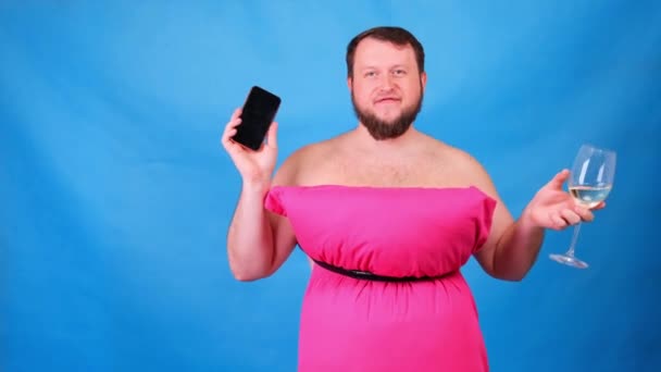 Tipul cu barbă amuzantă într-o rochie roz din perne cu un pahar de vin face selfie pe un fundal albastru. Carantină nebună. Curăţenie amuzantă. Modă 2020. Pune o pernă. Provocarea 2020 din cauza — Videoclip de stoc