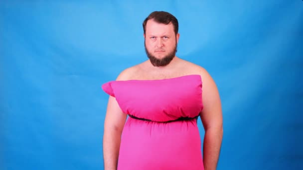 Grappige man met een baard in een roze jurk gemaakt van kussens is verdrietig op een blauwe achtergrond. Gekke quarantaine. Grappige schoonmaak. Mode 2020. Leg een kussen op. Uitdaging 2020 als gevolg van huisisolatie. — Stockvideo