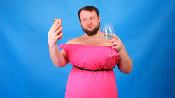 Cara barbudo engraçado em um vestido rosa feito de travesseiros com um copo de vinho faz selfie em um fundo azul. Quarentena maluca. Limpeza de casa engraçada. Moda 2020. Põe uma almofada. Desafio 2020 devido a — Fotografia de Stock