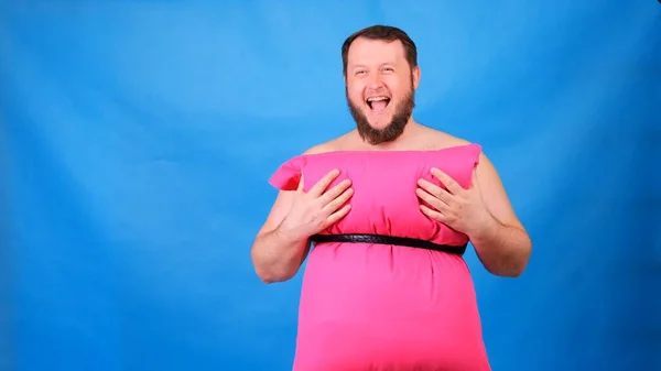Rolig skäggig kille i rosa klänning gjord av kuddar vidrör bröstet på en blå bakgrund. Galen karantän. Rolig städning. Mode 2020. Sätt på dig en kudde. Utmaning 2020 på grund av isolering — Stockfoto