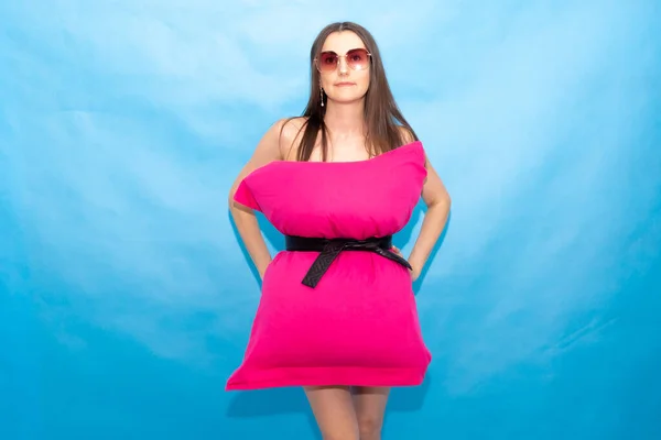 Söt kvinna i rosa kudde klänning och solglasögon på en blå bakgrund för galna kudde utmaning på grund av att stanna hemma isolering. Caronavirus-karantän. Modeflickan. Kudde samtal på grund av isolering i hemmet. — Stockfoto