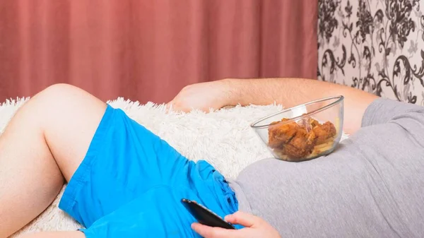 뚱뚱 한 남자는 TV 채널을 바꾸고 패스트 푸드치킨 날개를 먹는다. 집에서의 지루 함, 영양실조, 게으름의 개념. — 스톡 사진