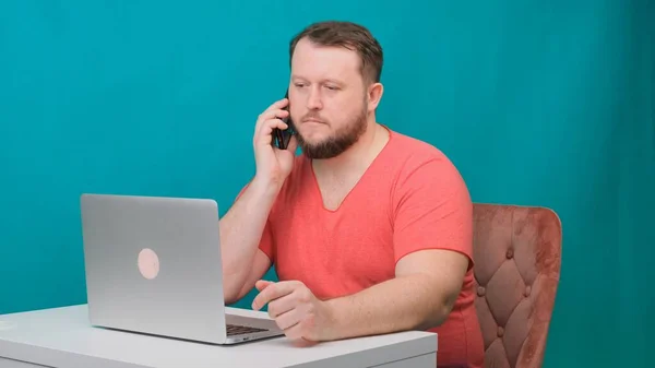 Giovane uomo d'affari felice in una t-shirt rosa sta lavorando su un computer portatile e parlando su un telefono cellulare su uno schermo verde. Ritratto di un uomo parlante che guarda nel suo portatile. L'uomo che lavora alla sua scrivania nel — Foto Stock