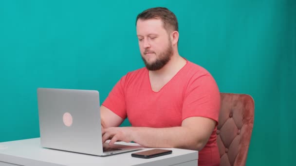 Il giovane uomo d'affari felice in una t-shirt rosa lavora su un computer portatile su uno schermo verde. Ritratto di un uomo parlante che guarda nel suo portatile. Uomo che lavora alla sua scrivania in ufficio. — Video Stock