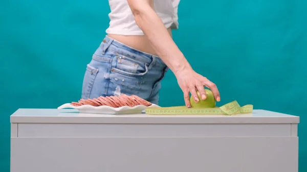 Frau wählt zwischen Apfel oder Wurst auf grünem Hintergrund. Das Konzept einer gesunden Ernährung, Ernährung und Willenskraft. — Stockfoto