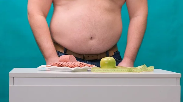 Mann mit dickem Bauch wählt zwischen Wurst und grünem Apfel. Das Konzept der Ernährung und Willenskraft. — Stockfoto