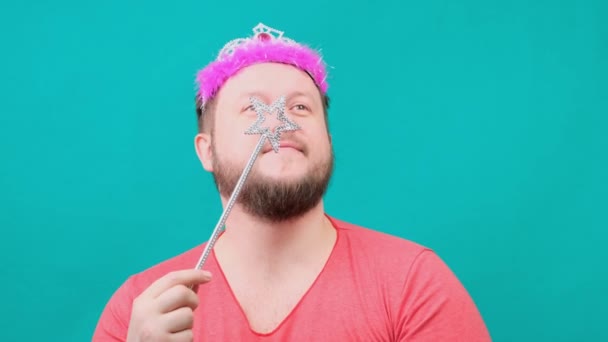 Lindo hombre barbudo raro en una camiseta rosa con un deadema en la cabeza sueña con una varita mágica en la mano. Un divertido chiste de mago para hacer y cumplir un deseo . — Vídeos de Stock