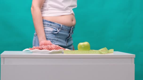 Mujer preocupada con un vientre gordo debe elegir entre una manzana verde y una salchicha. Concepto de dieta, hermosa mujer joven elegir entre comida saludable y comida chatarra — Vídeos de Stock