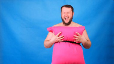 Yastıklardan yapılmış pembe elbiseli komik sakallı adam mavi arka planda göğsüne dokunuyor. Çılgın karantina. Garip ev temizliği. Moda 2020. Yastık koy. Ev izolasyonu nedeniyle 2020 Meydan Okuma