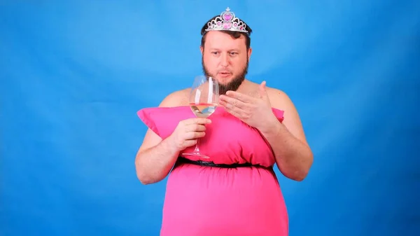 Lustiger bärtiger Typ in einem rosafarbenen Kleid aus Kissen in einer Krone mit einem Glas Wein auf blauem Hintergrund. Verrückte Quarantäne. Lustige Hausreinigung. Mode 2020. Auf ein Kissen legen. Herausforderung 2020 — Stockfoto