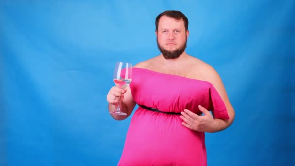 Grappige man met een baard in een roze jurk gemaakt van kussens drinkt wijn uit een glas op een blauwe achtergrond. Gekke quarantaine. Mode 2020. Leg een kussen op. Uitdaging 2020 als gevolg van huisisolatie — Stockvideo