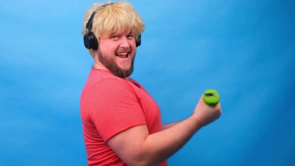 Divertente grasso uomo freaky in una parrucca con manubri facendo esercizi su uno sfondo blu. — Video Stock