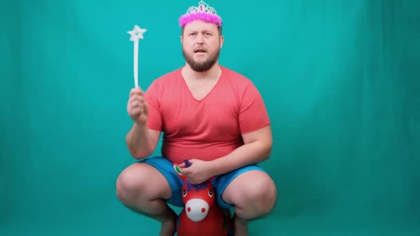 분홍색 티셔츠를 입고 마법 지팡이를 손에 든 유니콘을 타고 머리에 왕관을 쓰고 있는 수염을 기른 젊은 남자. 소원을 이루기 위한 웃긴 마법사의 농담. — 비디오