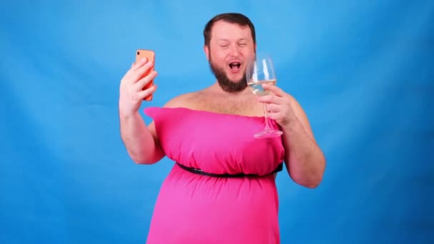Chico barbudo divertido en un vestido rosa hecho de almohadas con una copa de vino hace selfie sobre un fondo azul. Cuarentena loca. Una limpieza divertida de la casa. Moda 2020. Ponte una almohada. Reto 2020 debido a — Vídeos de Stock