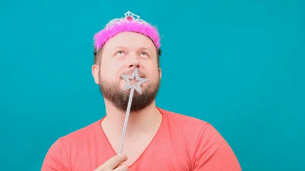 Divertido barbudo hombre raro en una camiseta rosa con un deadema en la cabeza está soñando con una varita mágica en la mano. Un divertido chiste de mago para hacer y cumplir un deseo . — Foto de Stock